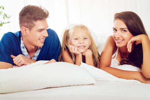 Beautiful young parents with their cute daughter lying in bed and looking at the camera with a smile.