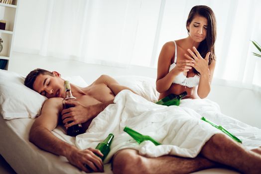 Young woman had just woke up next to a naked man who sleeps next to pile of empty bottles with disappointment and sadness looking at the wedding ring on her hand.