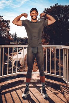 Young urban smiling sportsman pointing his muscle after training at the bridge and looking at camera.