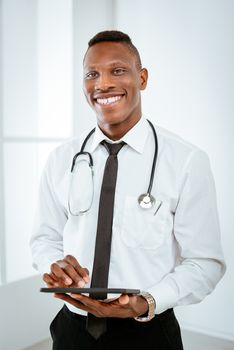 African smiling doctor with stethoscope standing in the doctor's room and using digital tablet.