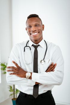 African smiling doctor with stethoscope standing with arms crossed in the doctor's room and looking at camera.