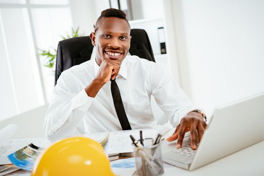 Smiling African construction engineer sitting at desk in the office and looking at camera. 