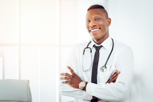 African smiling doctor with stethoscope standing with arms crossed in the doctor's room and looking at camera.