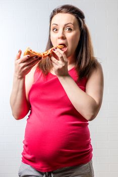 Hungry pregnant woman with a large slice of pizza on a brick white wall background