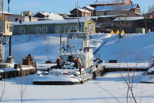 The frozen river port. Cranes, barges, old buildings. An ice-covered river covered with snow and a dry forest in the background