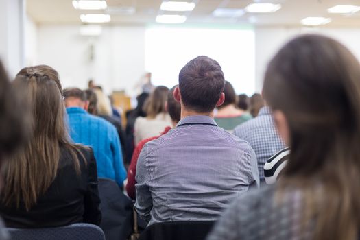 Business and entrepreneurship symposium. Female speaker giving a talk at business meeting. Audience in conference hall. Rear view of unrecognized participant in audience. Copy space on whitescreen.