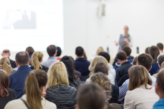 Business and entrepreneurship symposium. Female speaker giving a talk at business meeting. Audience in conference hall. Rear view of unrecognized participant in audience. Copy space on whitescreen.