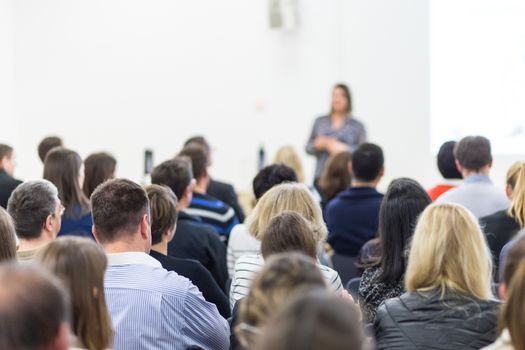 Business and entrepreneurship symposium. Female speaker giving a talk at business meeting. Audience in conference hall. Rear view of unrecognized participant in audience. Copy space on whitescreen.