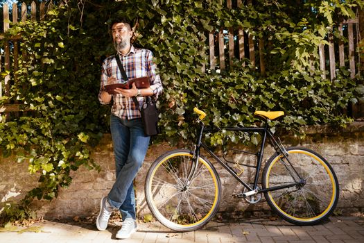 Pensive casual businessman taking his work to greener spaces. He is standing against the wall next to bike.