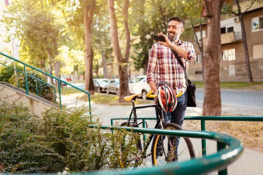 Casual businessman going to work by bicycle. He is walking next to bike, holding smartphone and waving Hello.