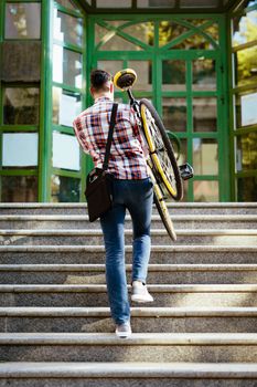 Casual businessman going to work by bicycle. He is carrying the bike upstairs to the entrance of the office building. Rear view.