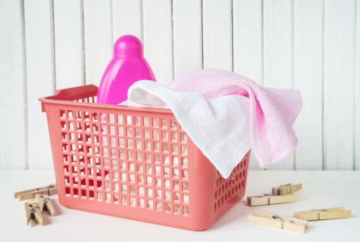 White and pink towels and detergent in pink bottle are in a red plastic laundry basket on the white wooden background