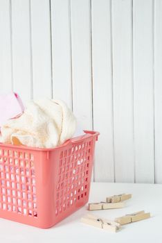 White, beige and pink towels in a red plastic laundry basket and wooden clothespins are on the white wooden background