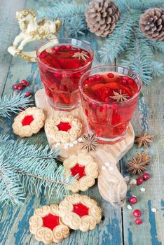 Christmas table: two glasses of red hot mulled wine with spices and cowberries and cookies with jam, surrounded by fir branches and cones