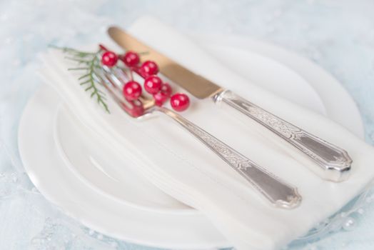 Silver knife and fork, and red holly berries and green thuja branches lie on the white porcelain plate