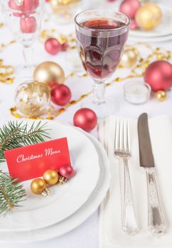Silver knife and fork, christmas ball and green spruce branche lie on the white porcelain plate, glass of red wine, which is located on a table covered with a white tablecloth