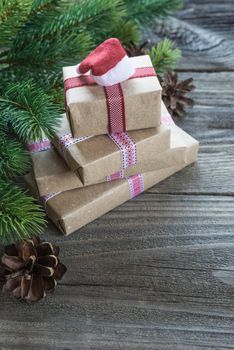 Christmas composition of pine cones, spruce branches and stack of gift boxes with Santa's cap on the background of old unpainted wooden boards; with copy-space