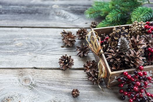 Christmas decoration: full wooden box of pine cones and red holly berries and spruce branches on the background of old unpainted wooden boards; Christmas composition for greeting card, websites, social media, business owners, magazines,  bloggers, artists etc.