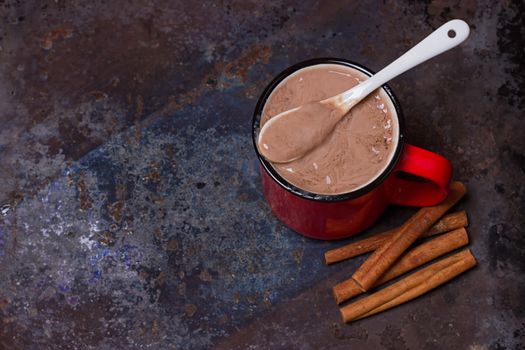 Vintage mug of hot chocolate with cinnamon sticks over dark background.