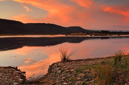 Lovely vibrant sunset colouring the clouds in reds and yellows and there hues cast onto the stony landscape and refletions  in the water.  Location  Boorooberongal Lake Penrith