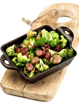 Homemade Useful Stew with Broccoli and Grilled Sausages in Black Iron Cast on Wooden Board on White background