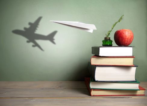 Paper plane with shadow of an aircraft next to a stack of books, quill and apple