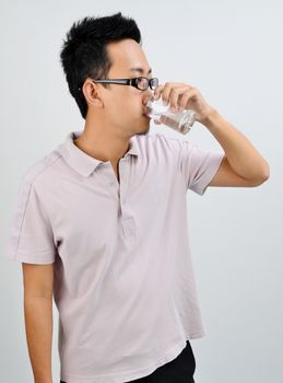 Portrait of Asian man drinking glass of mineral water, standing isolated on plain background.
