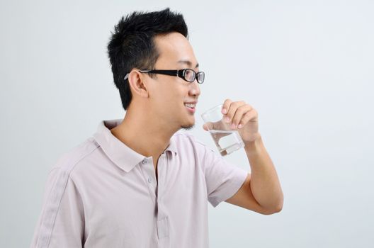 Portrait of Asian man smiling and drinking glass of mineral water, standing isolated on plain background.