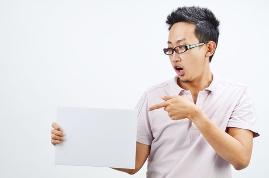 Portrait of shocked Asian man hand holding white blank paper card, standing isolated on plain background.