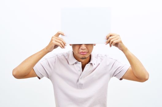 Portrait of sad Asian man hand holding white blank paper card covering his eyes, standing isolated on plain background.