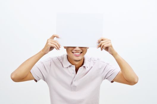 Portrait of Asian man hand holding white blank paper card covering his face, standing isolated on plain background.