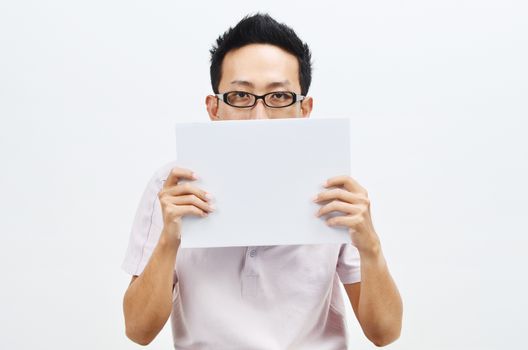 Portrait of sad Asian man hand holding white blank paper card covering his eyes, standing isolated on plain background.