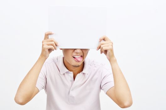 Portrait of Asian man tongue out and hand holding white blank paper card covering his eyes, standing isolated on plain background.