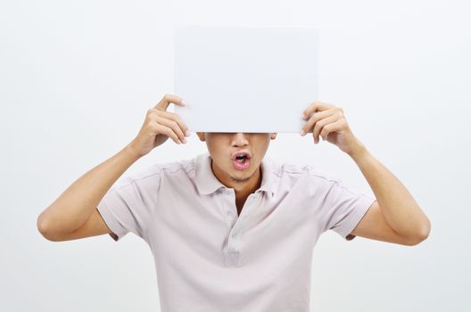 Portrait of shocked Asian man hand holding white blank paper card covering his eyes, standing isolated on plain background.