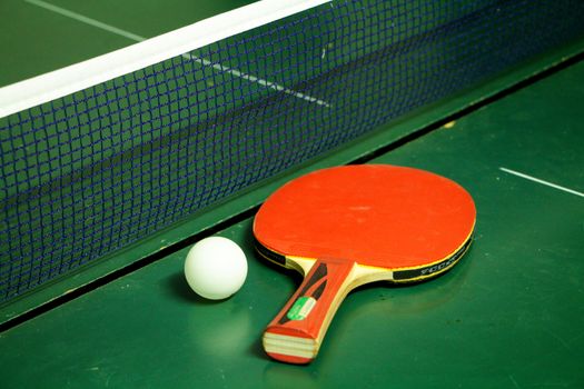 A red table tennis or ping pong racket and ball on a green table with blue net