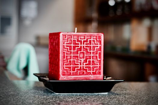 Big red square candle on the black plate with symetric pattern on front.