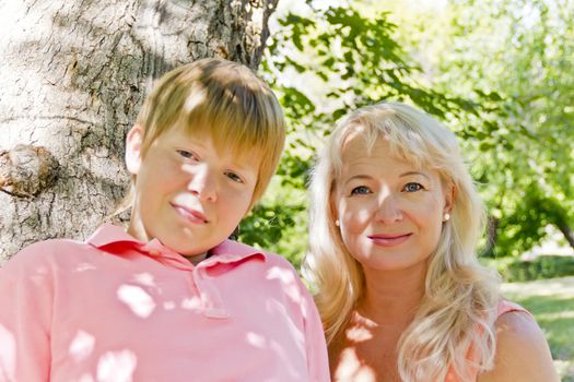 Blond woman and son smiling under green crone of tree in summer