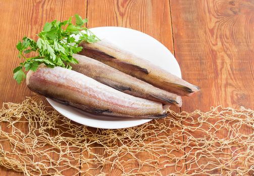 Uncooked carcasses of the Alaska pollock without of head and tail and bundle of parsley on a white dish on a surface of old wooden planks with fishing net
