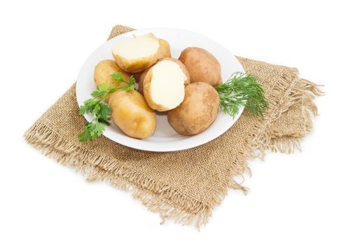 Two halves and several whole potatoes boiled in their jackets decorated with twigs of parsley and dill on the white dish on a sackcloth on a white background
