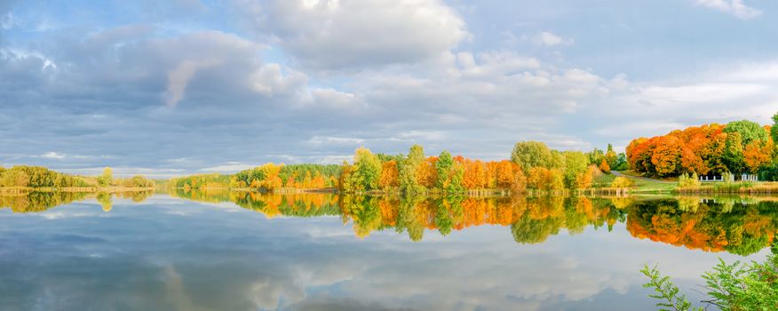 Panorama of the autumn forest and park on the shore of the lake, reflection of trees and sky with clouds in calm surface of the water
