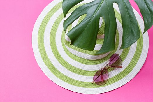 on green monstera leaf with sunglasses and straw hat on pink marble table