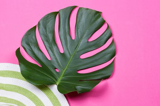 on green monstera leaf with sunglasses and straw hat on pink marble table