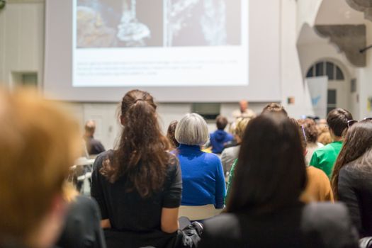 Male speaker giving presentation in lecture hall at university workshop. Audience in conference hall. Rear view of unrecognized participant in audience. Scientific conference event.