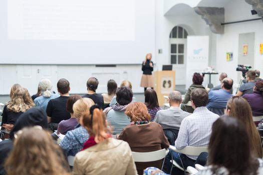 Business and entrepreneurship symposium. Female speaker giving a talk at business meeting. Audience in conference hall. Rear view of unrecognized participant in audience. Copy space on whitescreen.