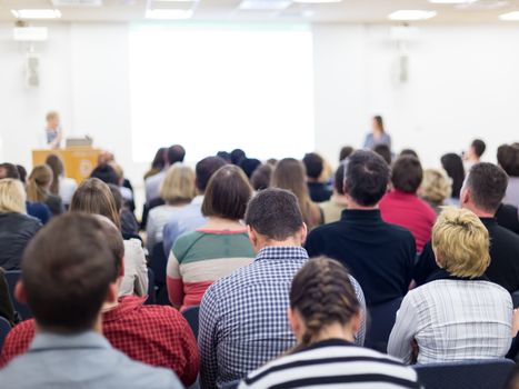 Business and entrepreneurship symposium. Female speaker giving a talk at business meeting. Audience in conference hall. Rear view of unrecognized participant in audience. Copy space on whitescreen.