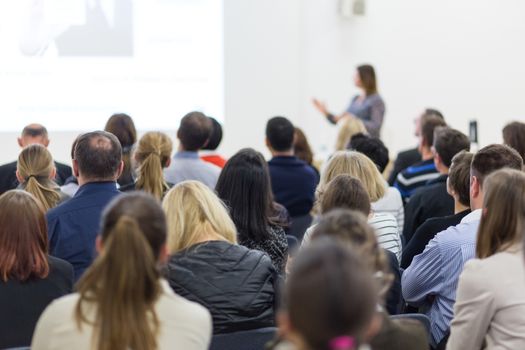 Business and entrepreneurship symposium. Female speaker giving a talk at business meeting. Audience in conference hall. Rear view of unrecognized participant in audience. Copy space on whitescreen.