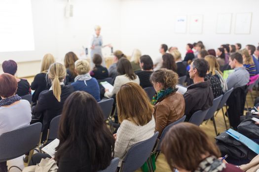 Business and entrepreneurship symposium. Female speaker giving a talk at business meeting. Audience in conference hall. Rear view of unrecognized participant in audience.