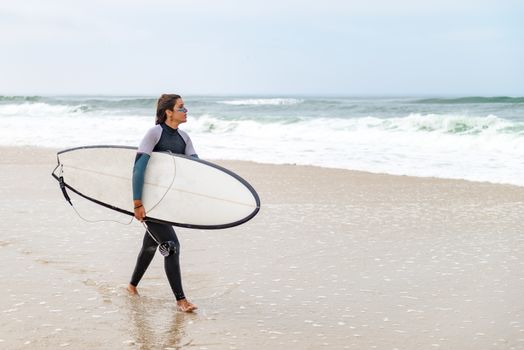 Young female surfer wearing wetsuit, holding surfboard under his arm, walking on beach after morning surfing session.