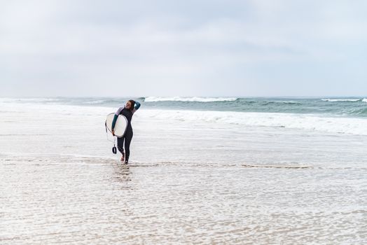 Young female surfer wearing wetsuit, holding surfboard under his arm, walking on beach after morning surfing session.