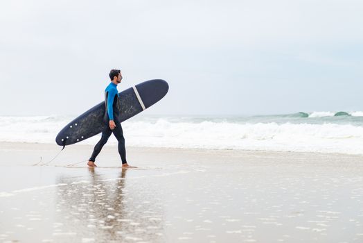 Young male surfer wearing wetsuit, holding surfboard under his arm, walking on beach after morning surfing session.